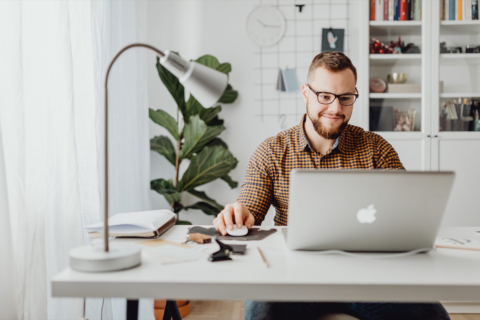 A man at a white desk using a silver laptop with a mouse beside a reading lamp